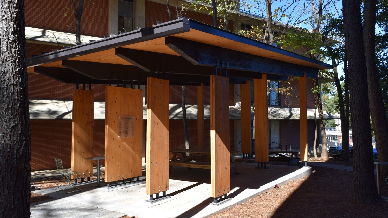 The Floating Grove, a wooden open-air structure with seating, is situated between Jordan Hall and Biltmore Hall at the College of Natural Resources, surrounded by trees and greenery.