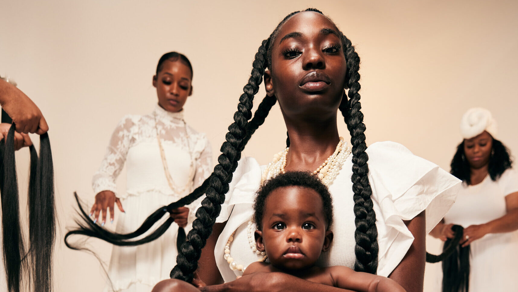 A Black woman with incredibly long braids holds a baby in a photo studio. Behind her stands two women who are holding and braiding the main subject's long hair.