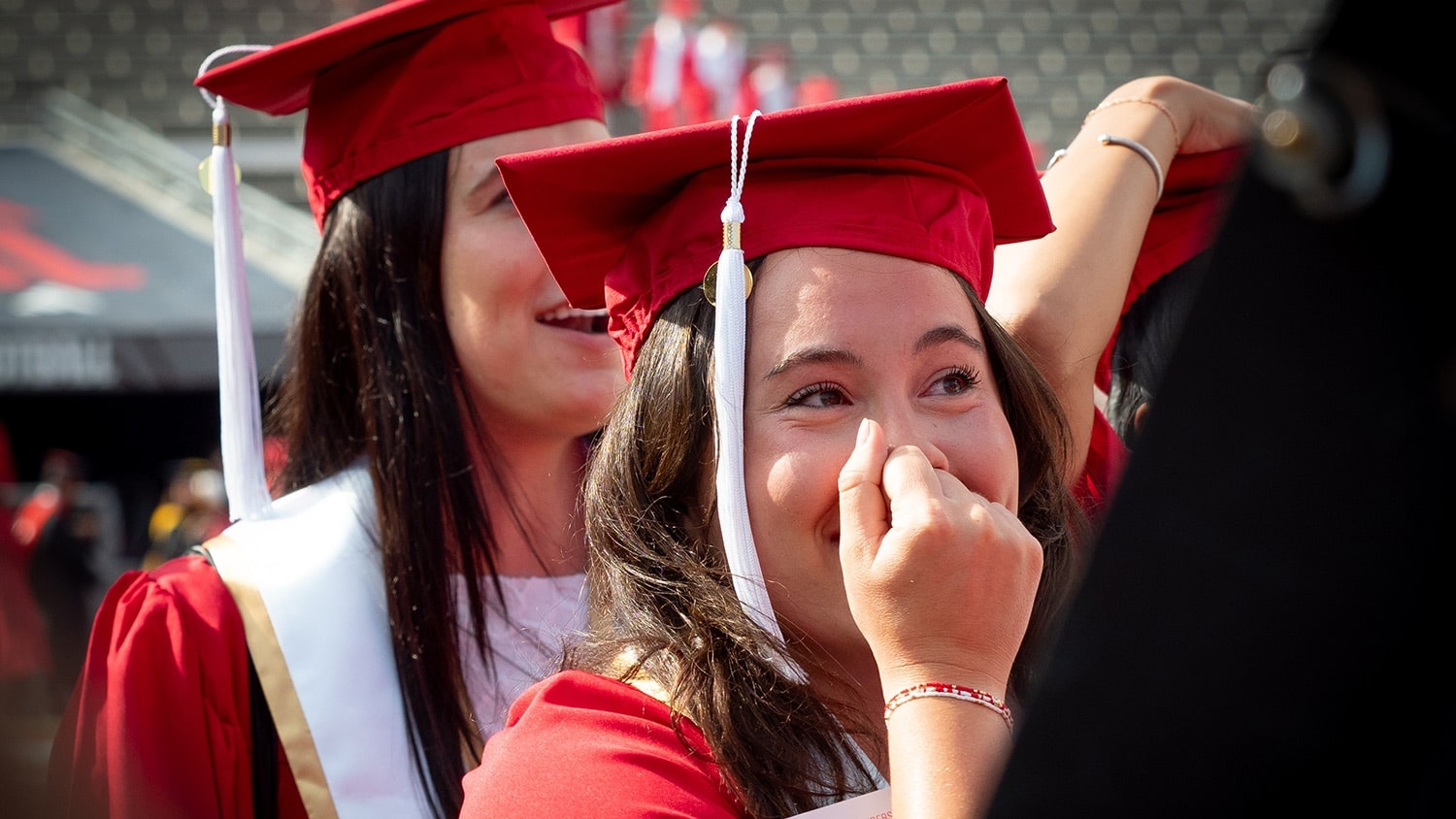 A student covers her mouth and smiles widely as she walks into commencement wearing her cap and gown.