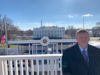 Ted Maliga poses in front of the presidential reviewing stand for the inauguration parade, with the White House in the background.